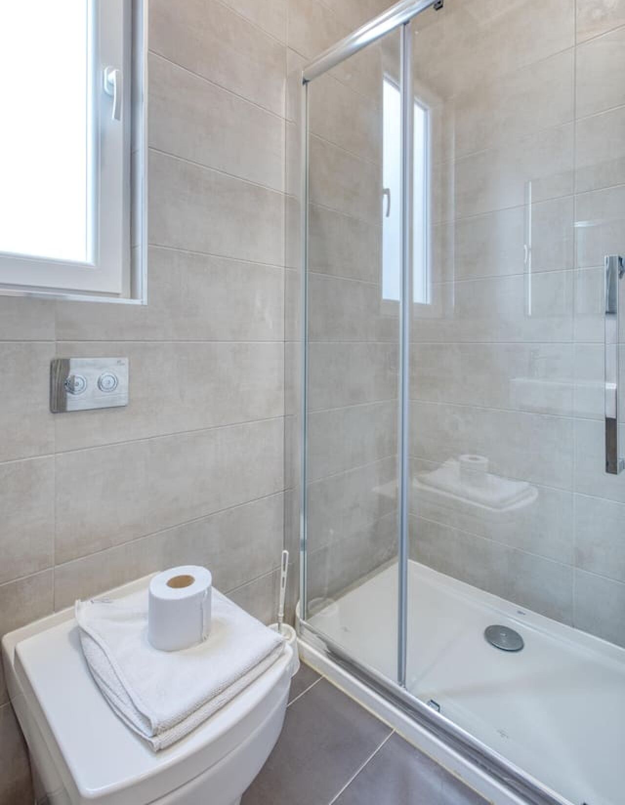 A modern bathroom with a glass shower enclosure, a toilet with a towel and toilet paper, and a window above the toilet.