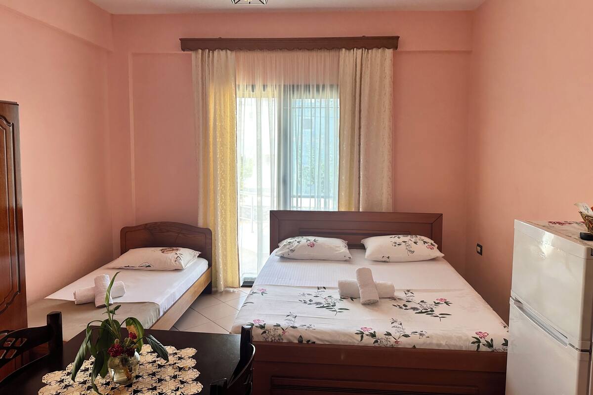 Hotellihuone, jossa on kaksi sänkyä, puinen vaatekaappi, pöytä maljakko täynnä kukkia ja jääkaappi.