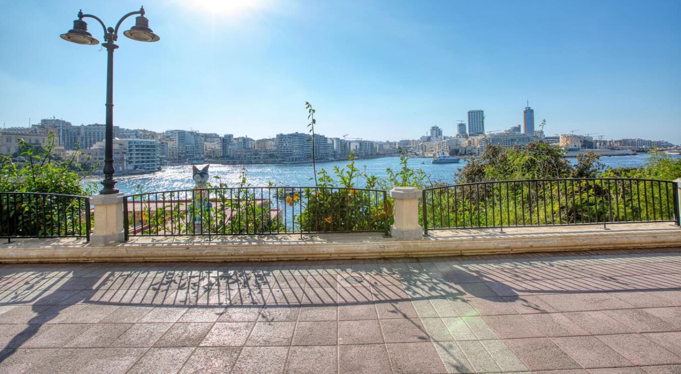 Eine Uferpromenade mit Metallgeländer, einer Laterne und Blick auf eine Stadtlandschaft über dem Wasser.