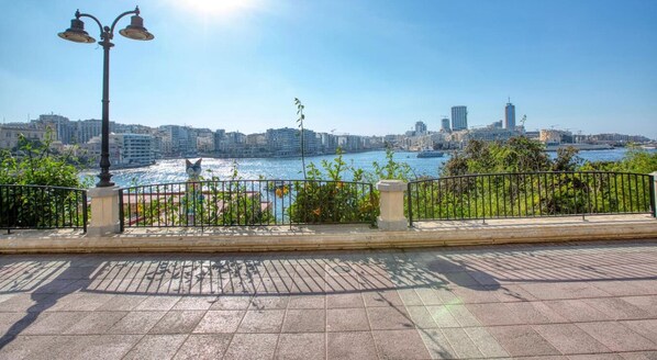 Une promenade en bord de mer, agrémentée d’une rambarde en métal, d’un lampadaire et offrant une vue sur les gratte-ciel d’une ville situés de l’autre côté de l’étendue d’eau.