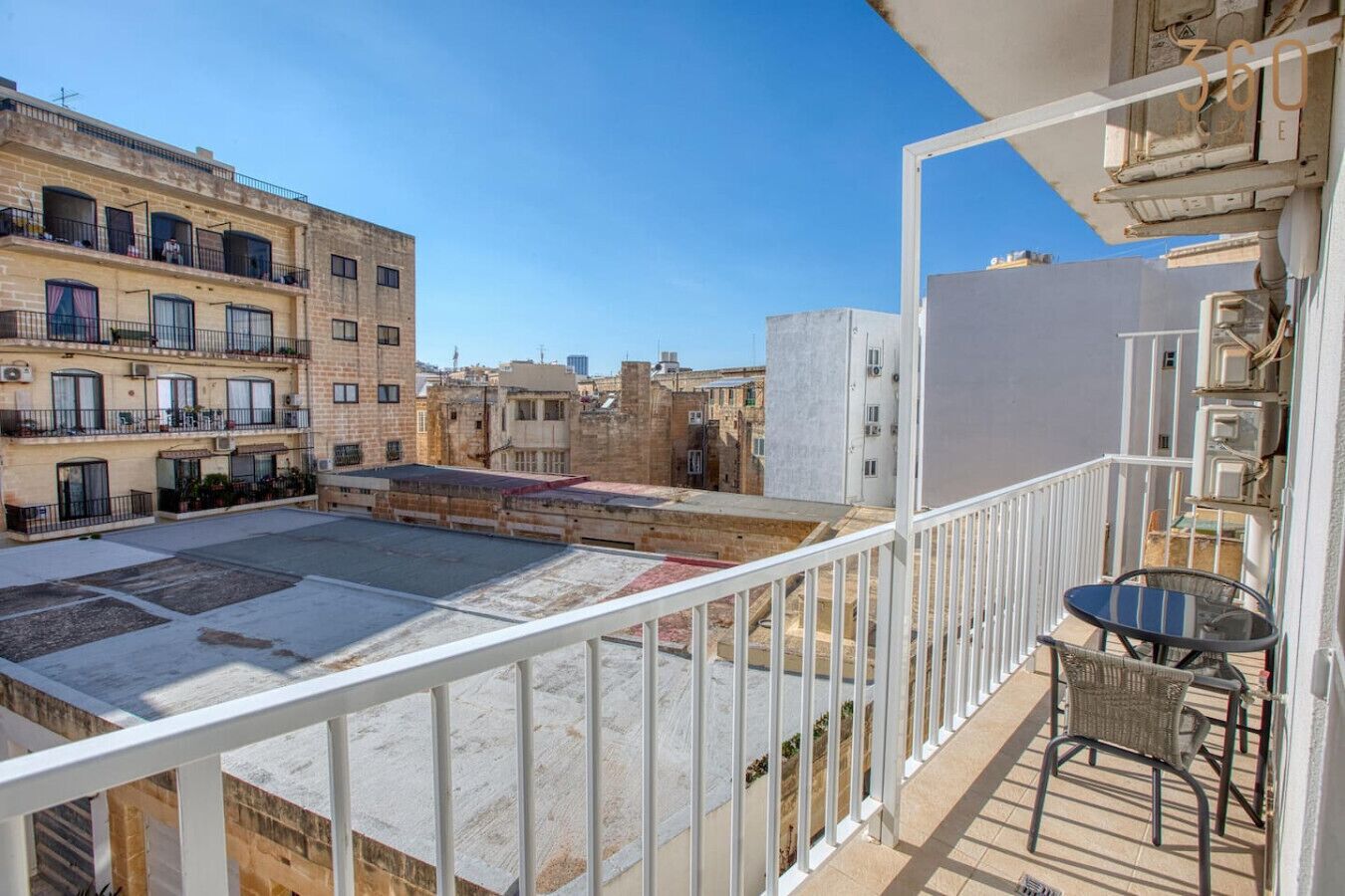 Una terraza en la azotea con barandilla blanca, una pequeña mesa con sillas y vistas de edificios circundantes.