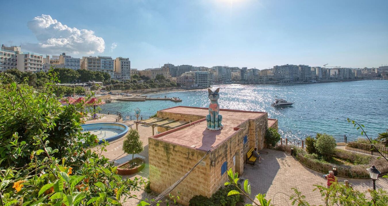 A coastal view with a building featuring a statue, a swimming pool, and a waterfront promenade.