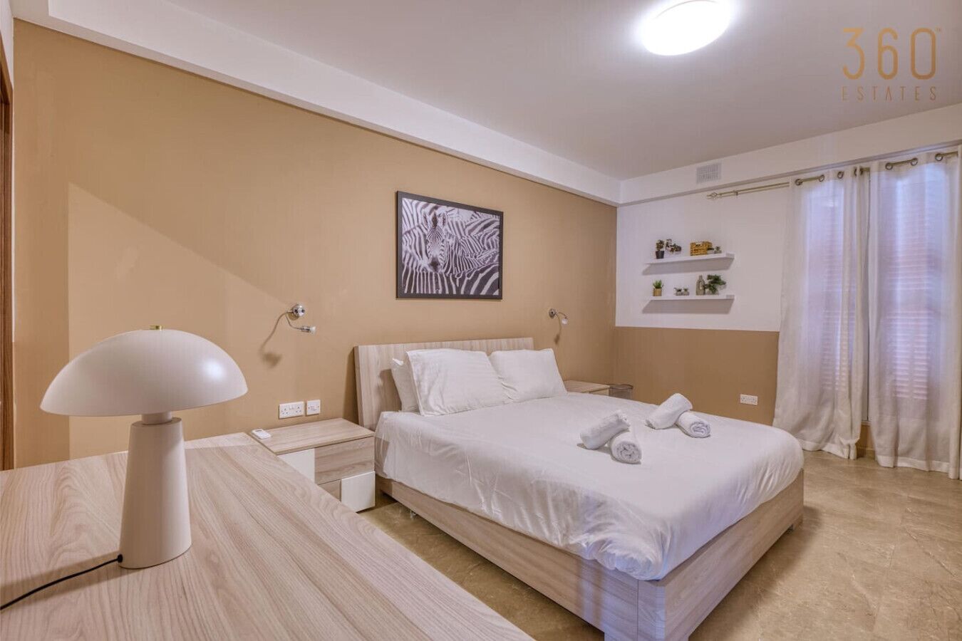 Habitación de hotel con cama, mesitas de noche, lámpara, estante en la pared con artículos de aseo y ventana con cortinas.