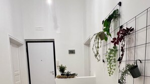 Ein Flur mit schwarzem Stuhl, einem kleinen runden Tisch und Kübelpflanzen an einer Gitterwand.