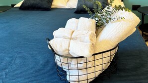 Ein ordentlich bezogenes Bett mit blauer Bettdecke, zwei Kissen und einem Metallkorb voller gefalteter Handtücher.