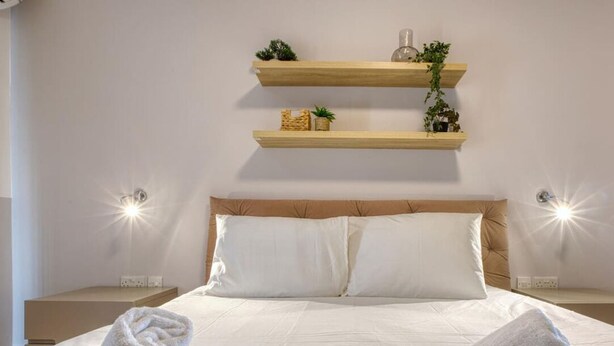 A neatly made bed with white linens and towels, wooden headboard, two bedside tables, and wall-mounted shelves with plants and decorative items. - Sunlit Sliema 2BR Penthouse, Steps to Seafront! (Sliema)