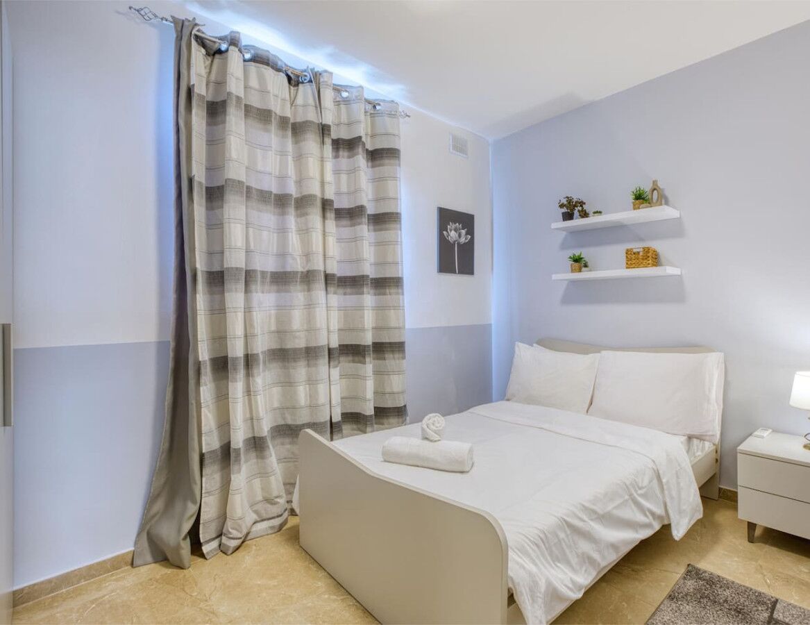 Un dormitorio con cama, mesita de noche, lámpara, repisa con adornos y cortina a rayas.