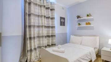Un dormitorio con cama, mesita de noche, lámpara, repisa con adornos y cortina a rayas.