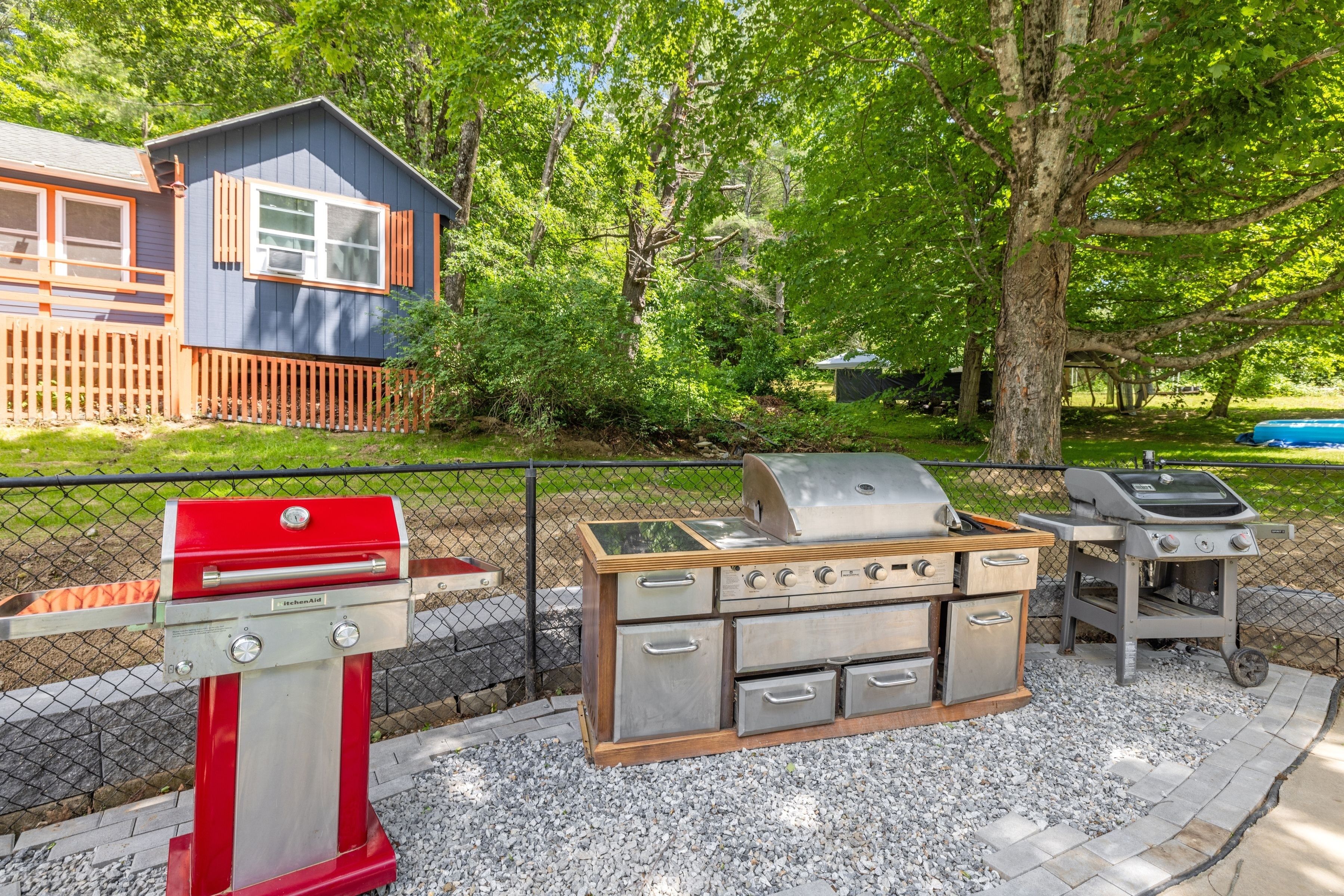 Ein Grillbereich im Freien mit einem roten Grill, einem Edelstahlgrill und einem Holzkohlegrill, umgeben von einem Zaun und Bäumen.