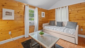 A cozy room with wooden walls, a sofa with patterned cushions, a glass coffee table with a plant, and a window with curtains.