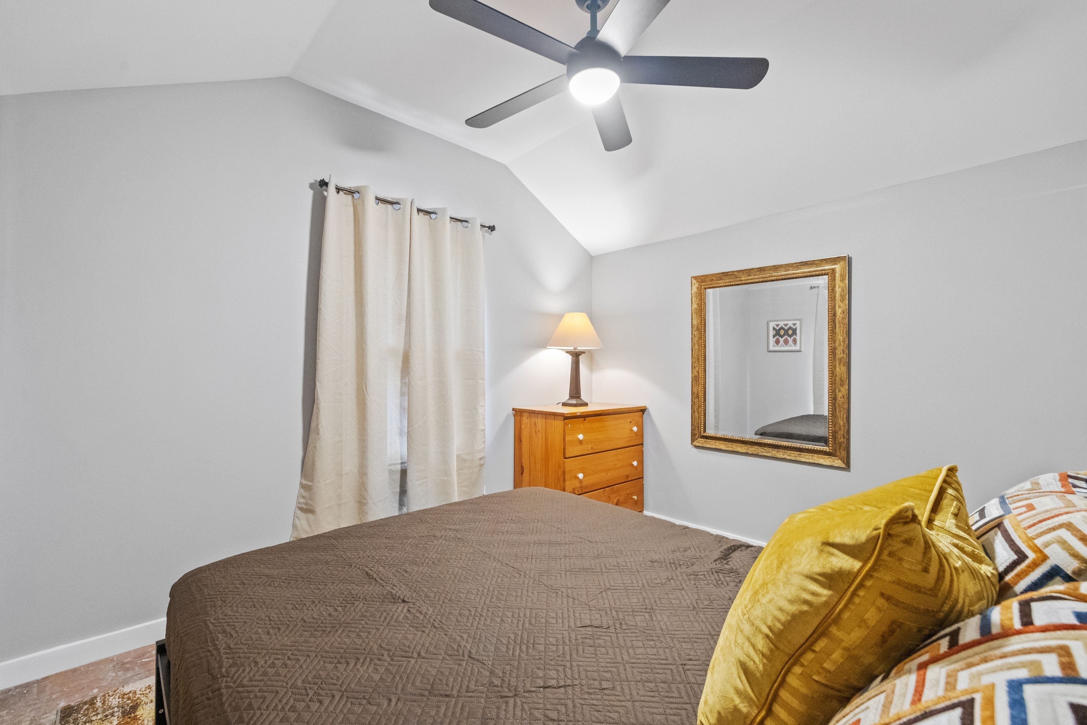Een slaapkamer met een bed, een plafondventilator, een houten dressoir, een lamp, een spiegel en een raam met gordijnen.