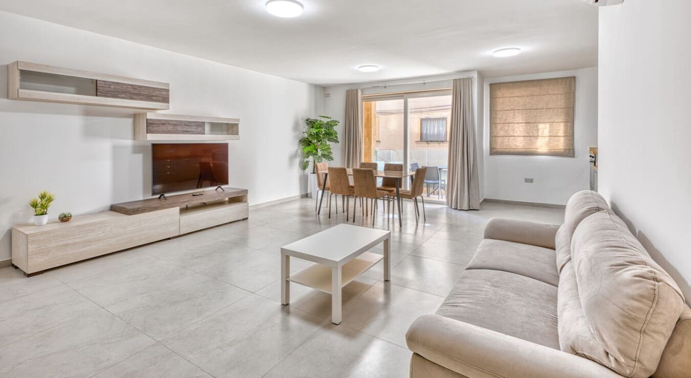 A modern living room with a sofa, TV, coffee table, and a dining area with chairs and a plant.