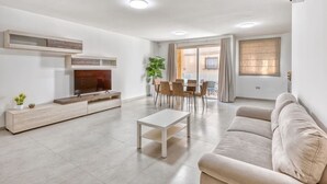 Ein modernes Wohnzimmer mit Sofa, Fernseher, Couchtisch und einem Essbereich mit Stühlen und einer Pflanze.