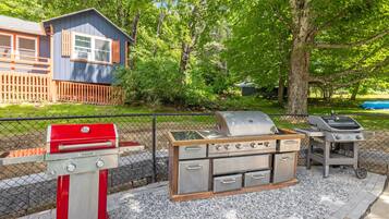 Area barbecue esterna con griglia rossa, griglia in acciaio inossidabile e griglia a carbone, circondata da una recinzione e alberi.
