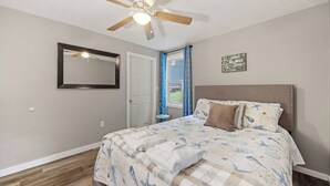 Un dormitorio con cama, ventilador de techo, puerta, ventana con cortinas y un cartel en la pared.