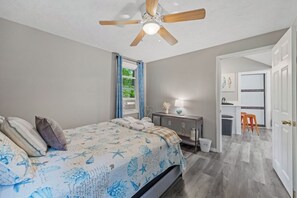 Een slaapkamer met een bed, een plafondventilator, een nachtkastje en uitzicht op een keuken.
