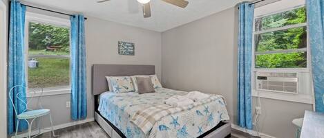 A bedroom with a bed, a ceiling fan, a window with curtains, a chair, and a sign on the wall.