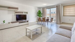 A modern living room with a flat-screen TV, a white coffee table, a beige sofa, and a dining area with a table and chairs.