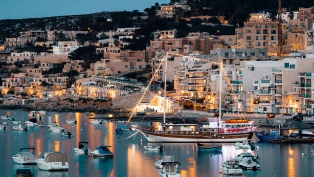 A coastal town at dusk with a large boat docked in the marina, surrounded by numerous smaller boats and buildings with lights on.