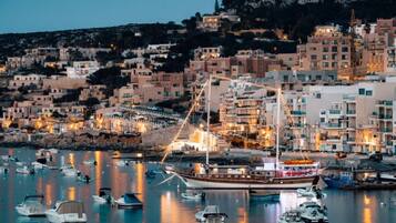A coastal town at dusk with a large boat docked in the marina, surrounded by numerous smaller boats and buildings with lights on.