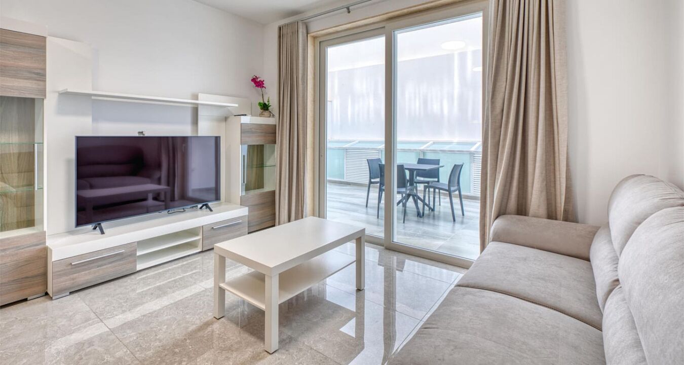A modern living room with a flat-screen TV, a sofa, a coffee table, and a balcony with outdoor seating.
