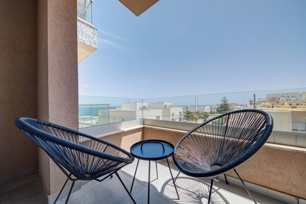 Un balcon avec deux chaises à l’armature en fil de fer noir et une petite table ronde, d’où l’on peut admirer le paysage urbain et la mer.