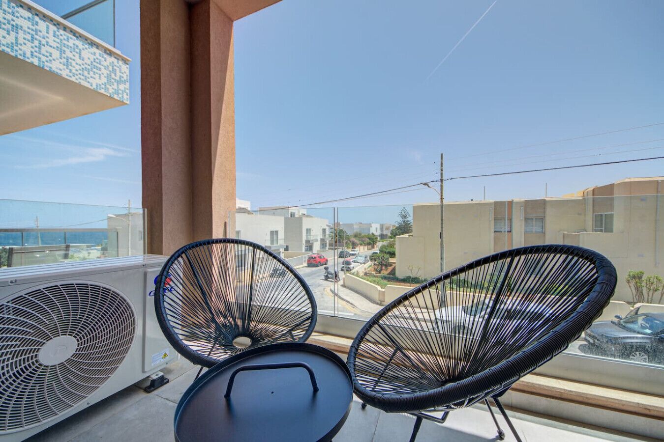 Un balcon avec deux fauteuils en osier noir et une table, donnant sur un quartier résidentiel avec des immeubles et des voitures garées.