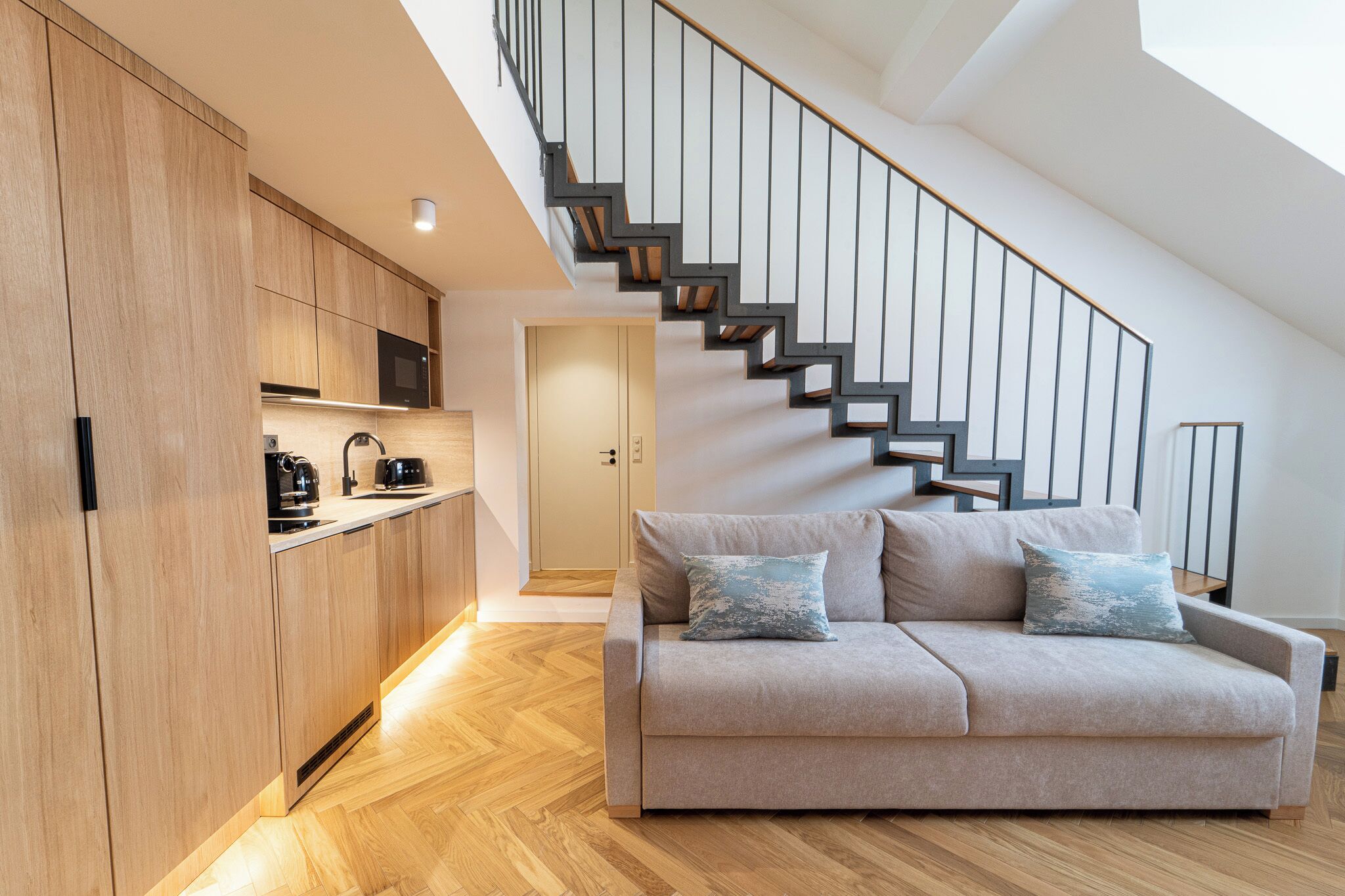 A modern kitchen with wooden cabinets, a built-in microwave, and a coffee maker. A staircase with black railings leads to a loft area.
