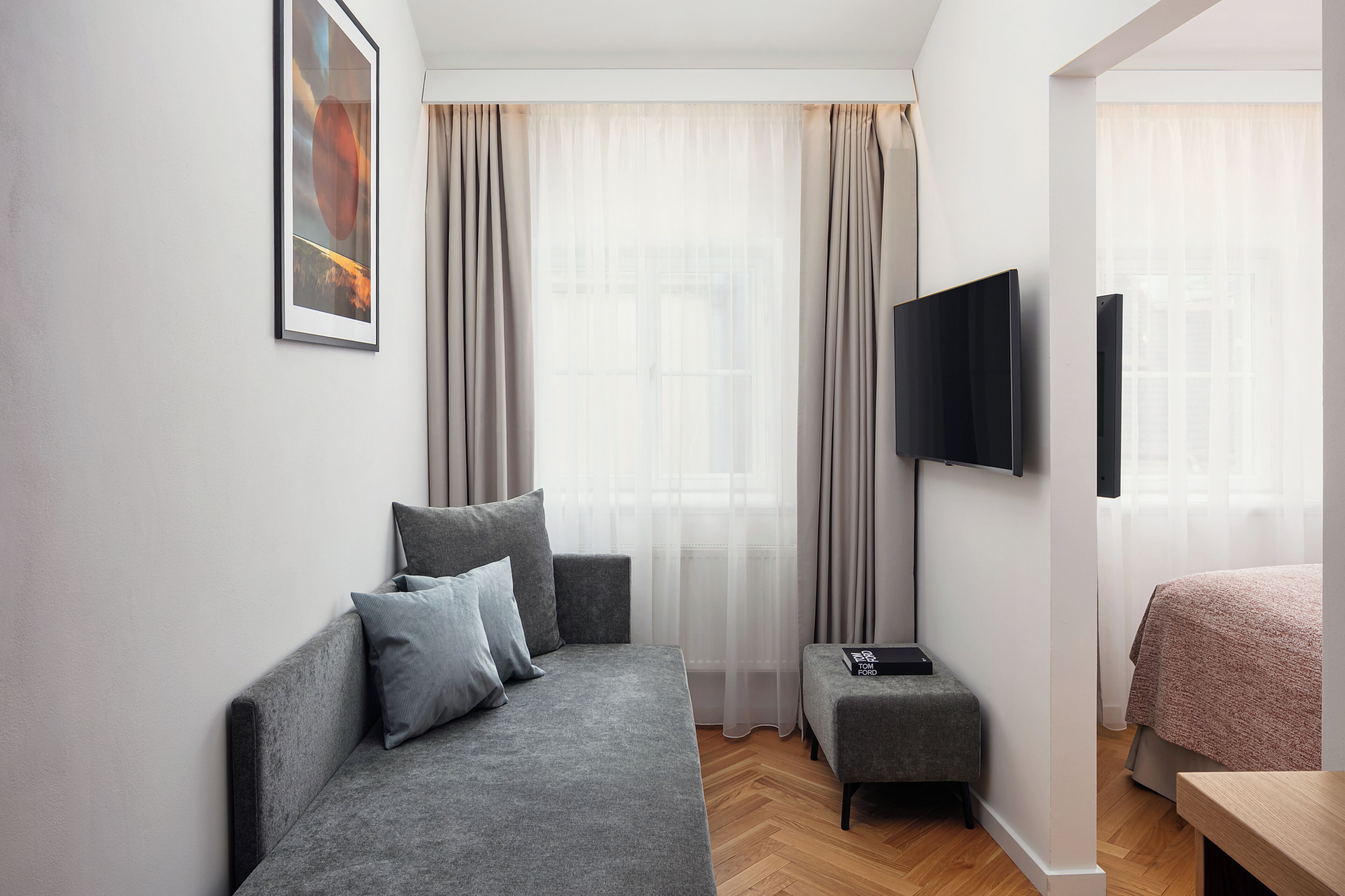 Una habitación de hotel moderna con un sofá gris, un pequeño reposapiés, un televisor de pantalla plana y una cama con una manta rosa.