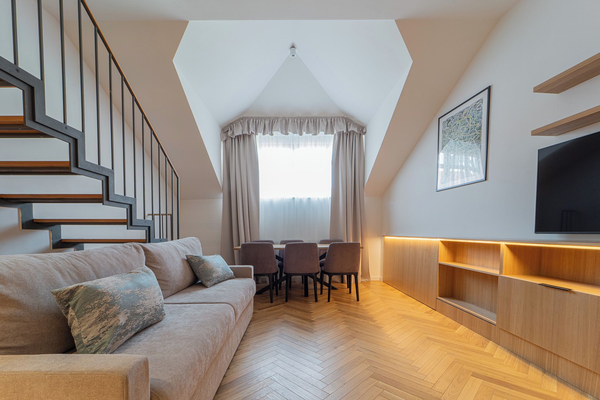 Una sala moderna con piso de espiga, un conjunto de sofá, una mesa de comedor con sillas, un televisor montado en la pared y una escalera con diseño geométrico.
