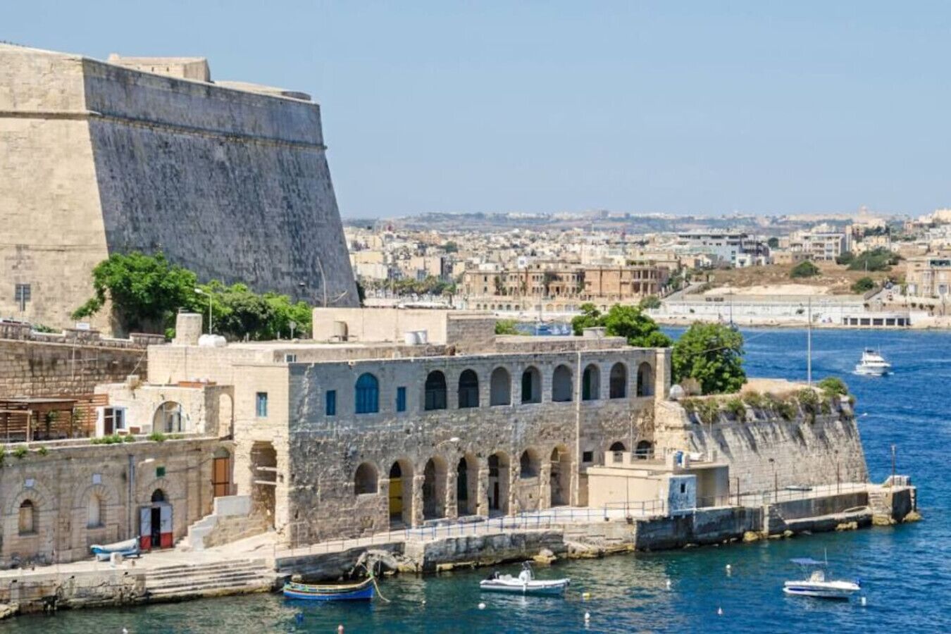 A coastal cityscape with a prominent fortress-like structure, a series of arches, and a marina with boats.