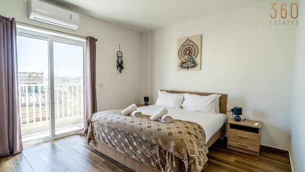 A modern hotel room with a large bed, a bedside table, a nightstand, a dreamcatcher, and a view of the city through the window.