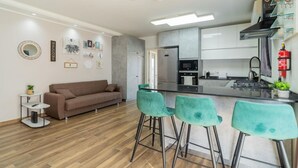 A modern kitchen with stainless steel appliances, a bar area with green bar stools, and a brown sofa in a living area.