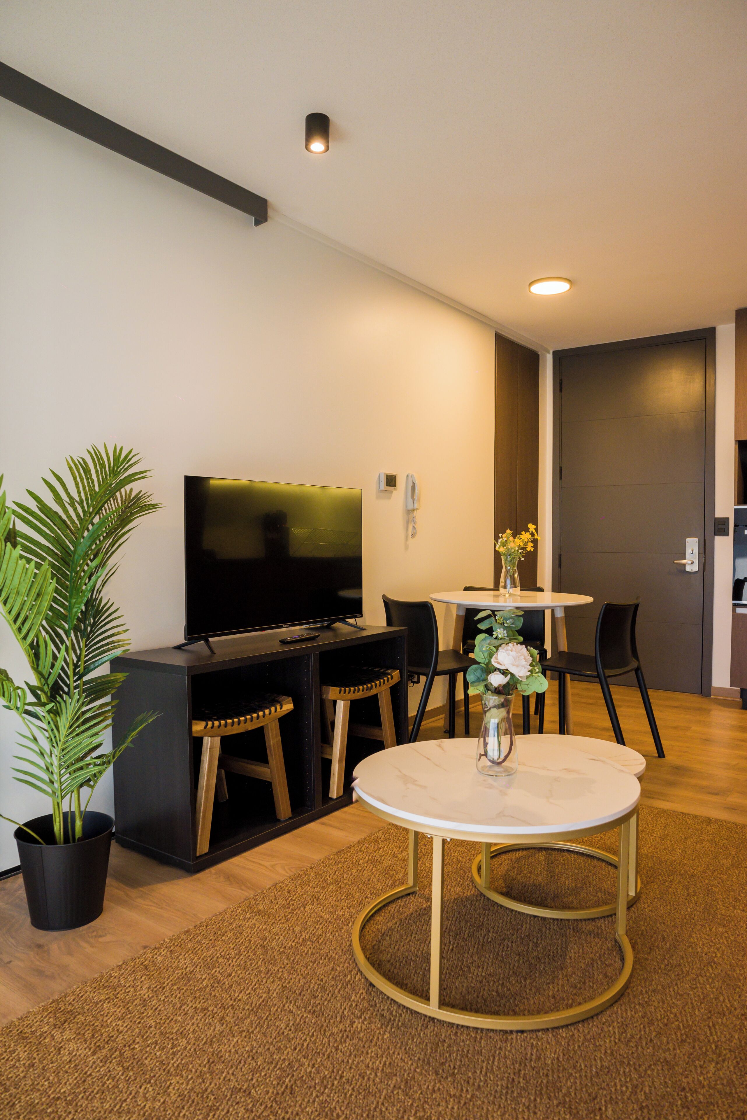 A modern living room with a flat-screen TV, a round coffee table with gold legs, two black chairs, a potted plant, and a vase with flowers.