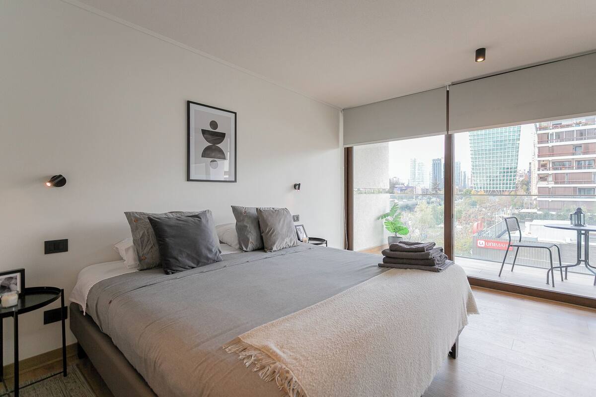 Ein modernes Schlafzimmer mit großem Bett, Blick auf die Stadt durch raumhohe Fenster und einem Balkon mit Gartenmöbeln.