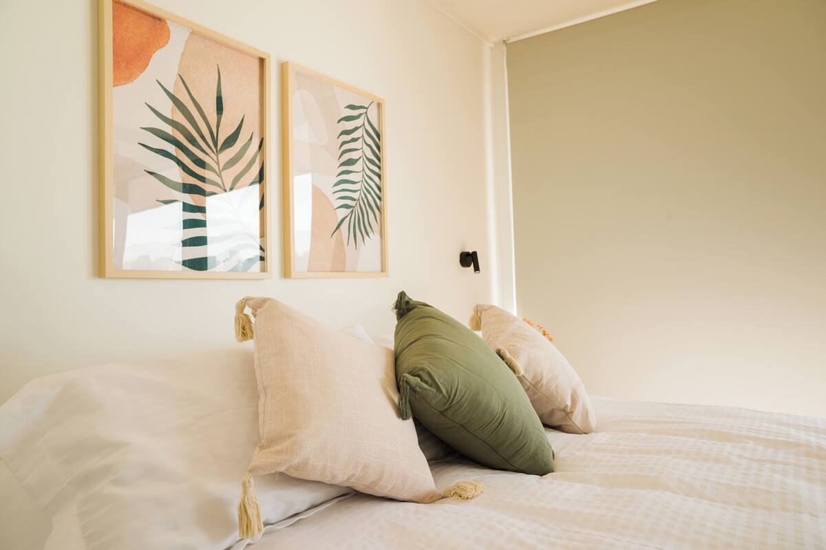 Ein Bett mit weißer Bettwäsche und zwei Kissen, eines grün und eines beige, in einem Zimmer mit schlichter Wand und einer Tür.