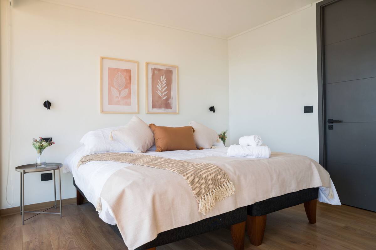 Ein ordentlich bezogenes Bett mit beiger Bettdecke und Kissen, ein kleiner Nachttisch mit Blumenvase sowie zwei gerahmte Blätterbilder an der Wand.