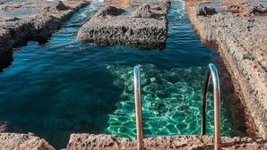Una costa rocosa con agua azul cristalina, una escalera metálica y veleros a lo lejos.