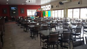 Un restaurante espacioso con numerosas mesas y sillas, un escenario con decoraciones coloridas, y grandes ventanales que dejan entrar luz natural.