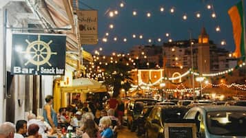 Une zone de restauration en plein air animée, avec des tables et des chaises, une enseigne indiquant un bar et un snack-bar, et une voiture bleue garée à proximité.