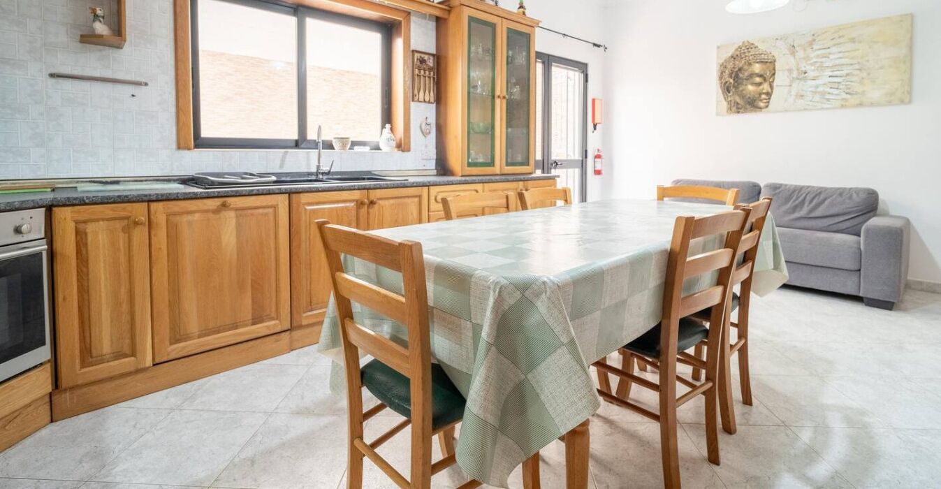 Uma cozinha com armários de madeira, uma mesa de jantar com cadeiras e um sofá cinza na sala de estar.