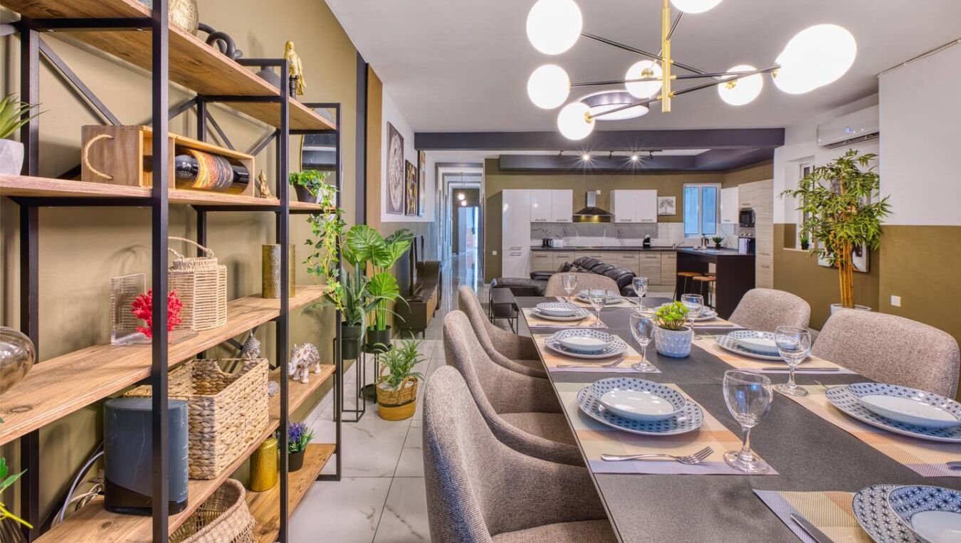 A modern dining area with a long table set for six, surrounded by comfortable chairs. There are shelves with decorative items and plants on the left.