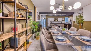A modern dining area with a long table set for six, surrounded by comfortable chairs. There are shelves with decorative items and plants on the left. - Lovely 3BR maisonette, spacious w/Wifi, AC & Pool (Swieqi)