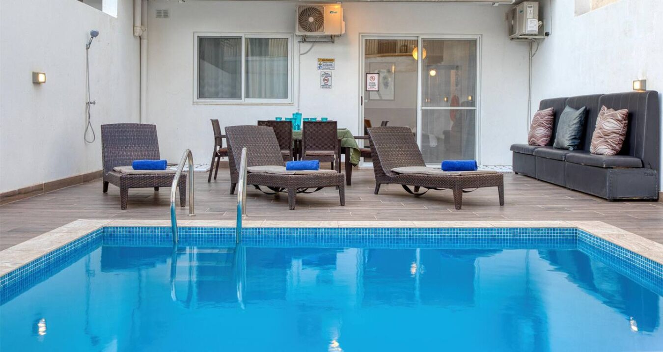 Ein Swimmingpool mit Liegestühlen und einer Sitzecke, eine Dusche und eine Tür, die zu einem Innenbereich führt.