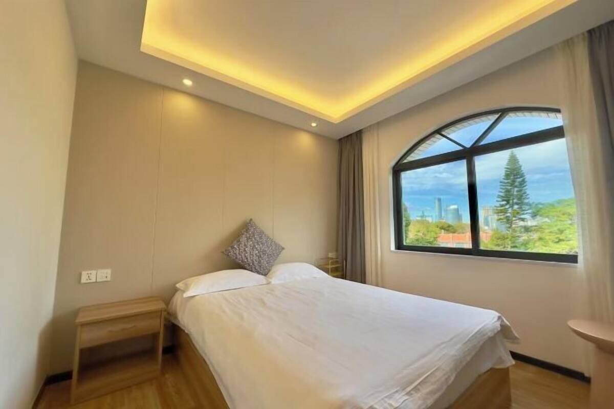 A bedroom with a bed, a nightstand, a window with a view, and recessed ceiling lighting.