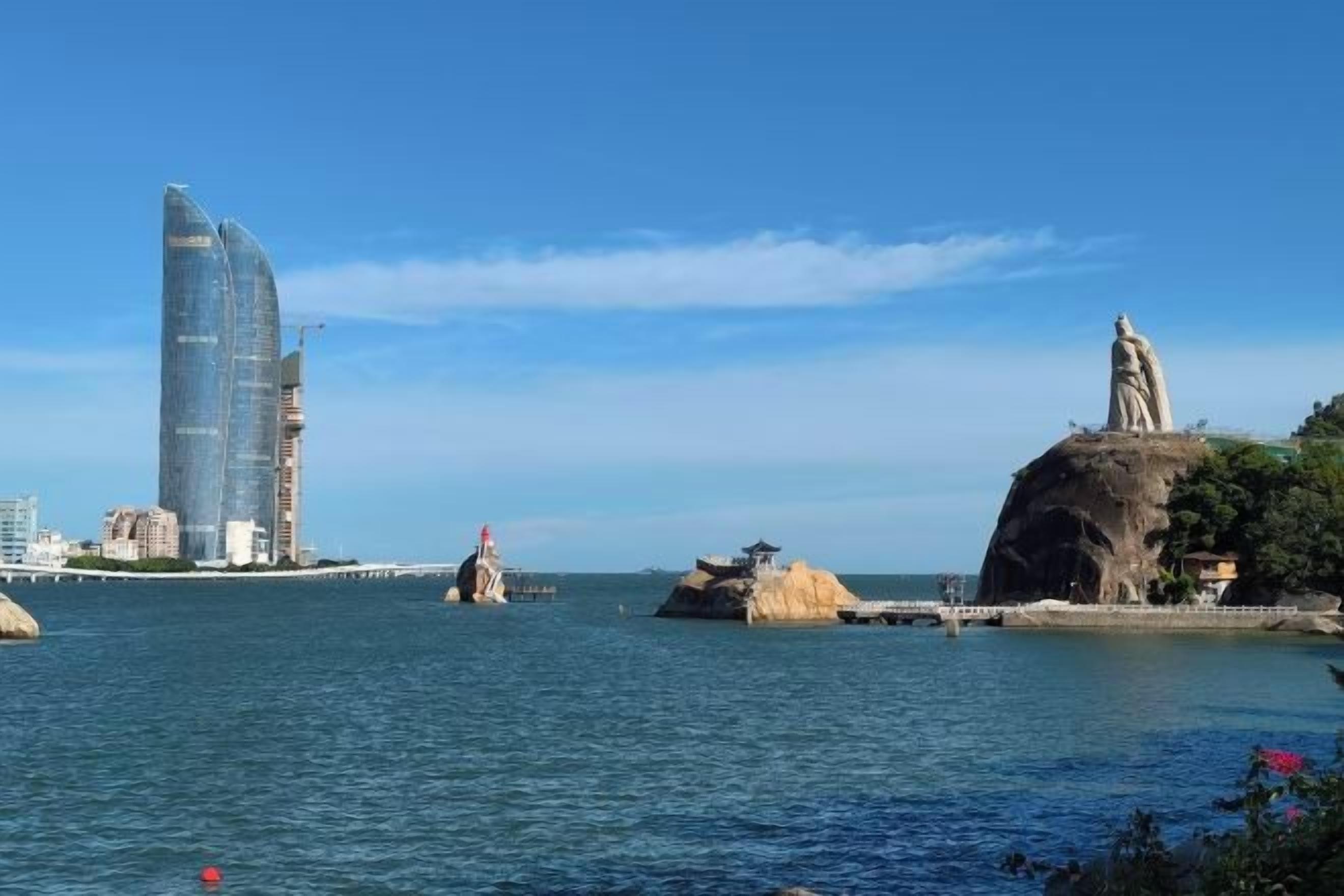 A coastal cityscape with a prominent modern skyscraper, a statue on a rock, and a bridge in the distance.