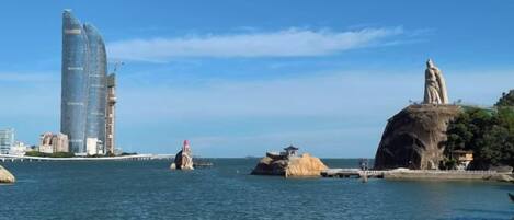 A coastal cityscape with a prominent modern skyscraper, a statue on a rock, and a bridge in the distance.