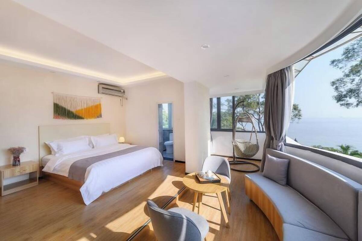 A modern hotel room with a large bed, a sofa, a small table, and a view of the sea.