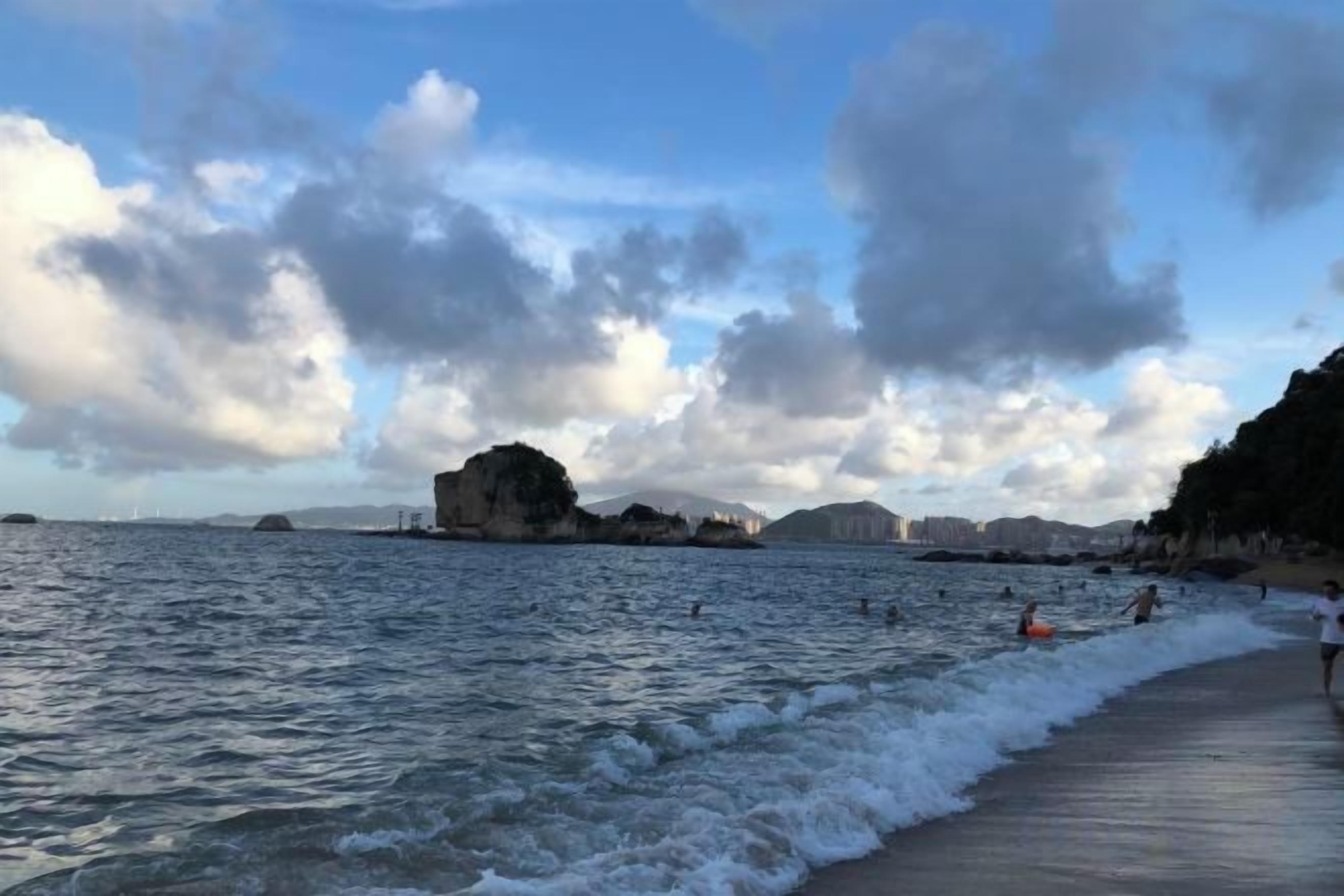Uma praia com ondas, um afloramento rochoso e edifícios ao longe.