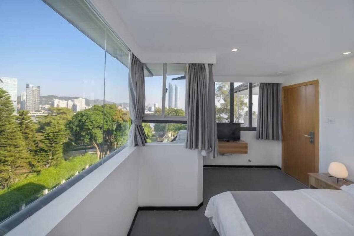 A modern hotel room with a large window, a bed, a TV, and a wooden door.